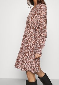 Brązowa sukienka w wzory z długimi rękawami i dekoltem w serek. Materiał zdobiony jest białymi abstrakcyjnymi kształtami, a sukienka ma luźny krój i rozszerzający się dół.