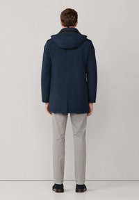 Marineblauer Kapuzenmantel mit elegantem Design, gefertigt aus einem glatten, wasserabweisenden Material. Begleitet von hellgrauen Hosen und dunklen Schuhen.