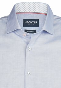 Lichtblauw overhemd met patroon en een moderne pasvorm. Voorzien van een button-down kraag en contrasterende, gepatterneerde binnenvoering. Twee knopen zichtbaar.