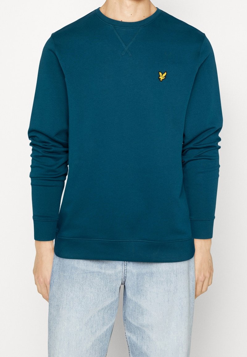 Sweatshirt teal en tissu doux, avec un col rond, des manches longues et un petit logo jaune brodé sur la poitrine.