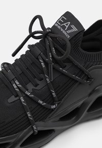 Schwarzer Sportschuh mit einem strukturierten Strick-Obermaterial, flachen Schnürsenkeln mit Logo-Akzenten und einer geformten Sohle mit einem markanten Design.