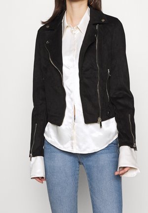 Personne portant une veste noire zippée sur une chemise en satin blanc et un jean bleu, debout devant un fond blanc uni.