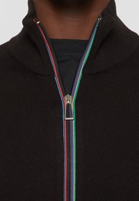 Svart zippered tröja med hög krage. Har flerfärgade dragkedjor i röd, blå, grön och gul mot en texturerad tyg.