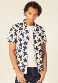 Lyhythihainen kudottu paita kermansävyisenä, jossa on tummansininen kukkakuosi. Ominaisuuksiin kuuluu napitus edessä ja rento istuvuus, yhdistetty valkoisen aluspaidan ja tummiin farkkuihin.