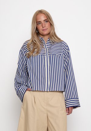 Mujer con cabello largo y rubio que lleva una camisa a rayas azul y blanca y pantalones beige de cintura alta, de pie con una mano en el bolsillo.