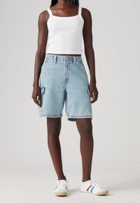 Persona vestida con una camiseta corta blanca, pantalones cortos de mezclilla cargo azul claros y zapatillas blancas con rayas rojas y azules, de pie contra un fondo liso.