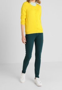 Žlutý kardigan s dlouhými rukávy, zapínání na knoflíky, kombinovaný s teplákovými úzkými kalhotami v barvě tyrkysu. Doplněno o bílé tenisky na neutrálním pozadí.