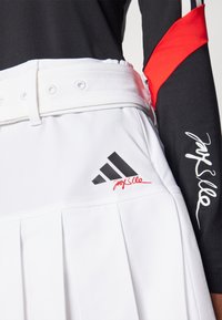 Λευκή πλισέ τενιστική φούστα με ελαστική μέση, που διαθέτει το μαύρο λογότυπο της Adidas και κόκκινη λεπτομέρεια υπογραφής. Μαύρο τοπ με κόκκινη λεπτομέρεια στο χέρι.