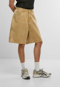 Bermudas de mezclilla beige con bolsillos laterales, presillas para cinturón y un corte relajado. Llevadas con zapatillas de correr plateadas y calcetines blancos hasta el tobillo; fondo sencillo.
