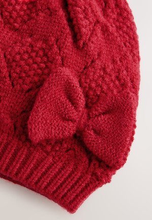 Rødt strikket hue med et tekstureret mønster, der har en stor, blød sløjfe som detalje. Ribbet kant nederst, der understreger det hyggelige design.