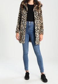 Kožich s leopardím vzorem s límcem z umělé kožešiny, doplněný modrými úzkými džínami s krajkovými detaily po stranách a černými teniskami.