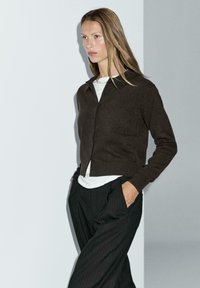 Brązowy sweter narzutowy z kołnierzykowym wzorem, zapięciem na guziki i ribbowymi mankietami, w połączeniu z białą koszulą i ciemnymi spodniami w paski.