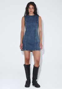 Farkkuliivitettu mekko, jossa on kiinteä siluetti, tummansininen väri, erottuva ompelu ja hieman liehuva helma. Mustat polvimittaiset saappaat on puettu jalkaan.