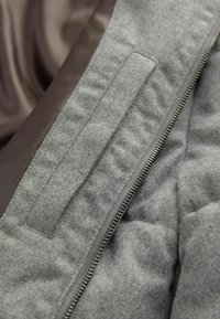 Tela de chaqueta acolchada gris con un forro marrón suave, con cremallera expuesta, panel gris texturizado y detalle de bolsillo.