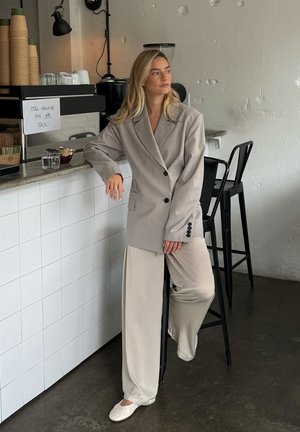 Woman in beige oversized blazer and wide pants leaning on a cafe counter with stacked cups and stools in a minimalist setting.