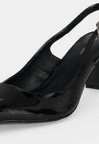 Γυναικεία slingback παπούτσια από μαύρο πατεντέ δέρμα με τετράγωνο τακούνι, μυτερή μύτη και ρυθμιζόμενο λουράκι με αγκράφα.