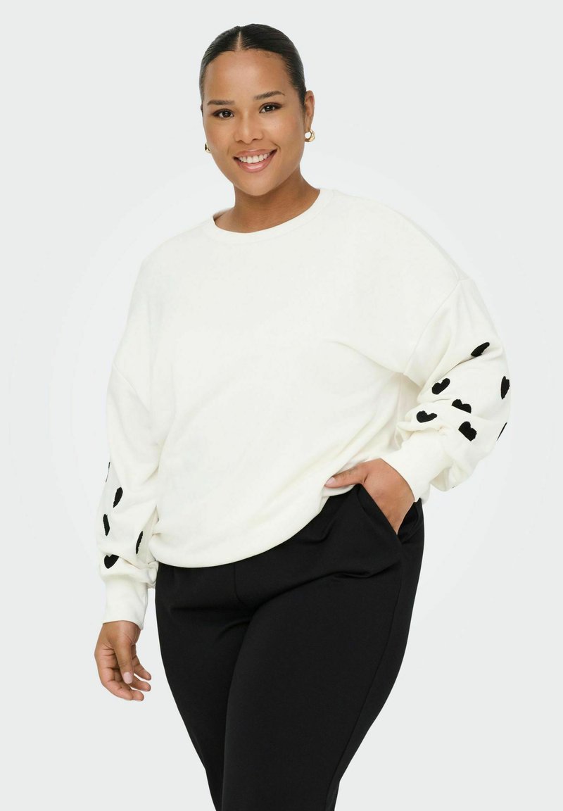 Sweatshirt blanc avec des motifs de cœurs noirs sur les manches, encolure ronde et coupe décontractée. Associé à un pantalon noir. Texture lisse.