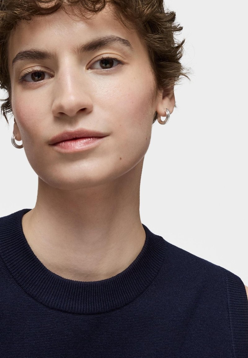 Suéter de manga corta en color navy con cuello redondo acanalado. El modelo tiene el pelo corto y rizado, y lleva pequeños aros de plata. Fondo neutro.