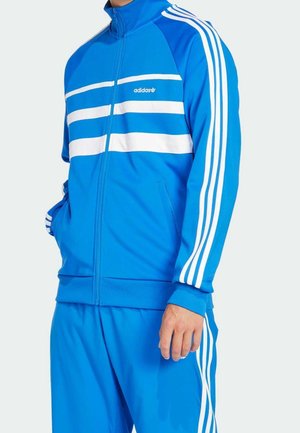 Mężczyzna w niebieskim dresie Adidas z białymi paskami na rękawach, klatce piersiowej i spodniach, z rękami w kieszeniach, stojący na jednolitym tle.