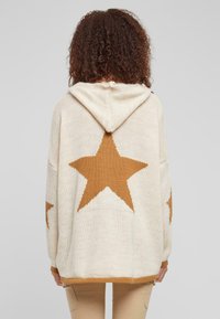 Cream-colored knitted hoodie with a large brown star pattern on the back and sleeves. Features a drawstring hood and ribbed hem.