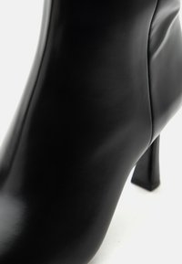 Gros plan d'une bottine noire en cuir à talon haut montrant une texture lisse et un design élégant sur fond blanc.