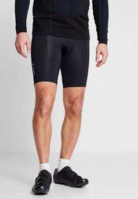 Shorts de cyclisme noirs avec une texture élastique et lisse ; dotés d'un rembourrage et d'une coupe ajustée. Portés avec des chaussures de cyclisme noires et des chaussettes blanches.