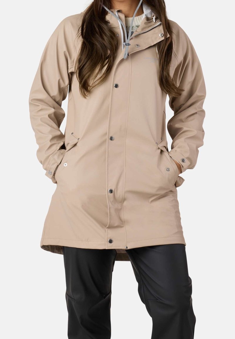 Femme portant un imperméable beige boutonné avec les mains dans les poches et un pantalon noir, montrée du cou au milieu des cuisses sur fond blanc.