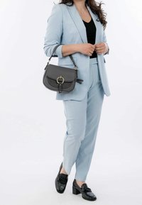 Hellblauer maßgeschneiderter Anzug mit einer taillierten Jacke und schmal zulaufenden Hosen, kombiniert mit einer dunkelgrauen Leder-Handtasche mit einem runden Akzent. Schwarze Slipper runden das Outfit ab.