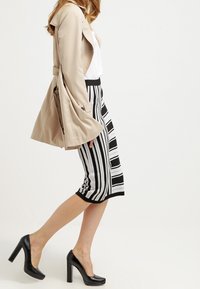 Beżowy płaszcz trencz na białej bluzce, zestawiony z czarno-białą spódnicą w paski i czarnymi szpilkami.