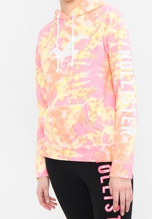 Personne portant un sweat à capuche tie-dye rose, jaune et orange avec "Hollister" sur la manche, associé à des leggings noirs ornés de texte rose le long d'une jambe.