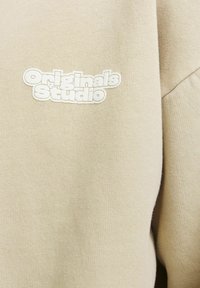 Beige sweatshirt featuring a textured fabric with an embroidered "Originals Studio" logo in white. Shows a close-up of the design detail.