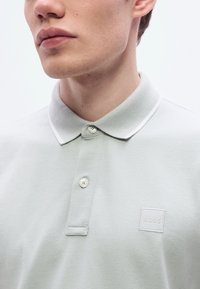 Ανοιχτό πράσινο πόλο μπλουζάκι με επίπεδη γιακά, δύο κουμπιά και μικρό λευκό λογότυπο. Η υφή του υφάσματος φαίνεται λεία και με υφ ή.