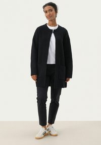Svart stickad cardigan med en texturerad mönster, öppen fram, två framfickor och långa ärmar, kombinerad med en vit skjorta och mörka byxor.