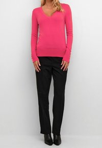 Pull ajusté à col en V de couleur rose vif avec des manches longues et des accents de boutons dorés aux poignets, associé à un pantalon noir ajusté et des bottes à talons.