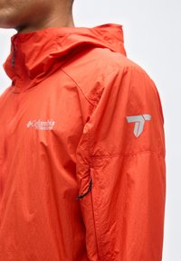 Gros plan sur une personne portant une veste Columbia Titanium orange vif avec capuche et poche zippée sur la manche, sur un fond uni.