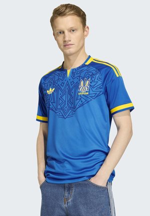 Junger Mann, der ein blau-gelbes ukrainisches Fußballtrikot mit geometrischen Mustern trägt, mit den Händen in den Taschen vor einem einfarbigen Hintergrund.