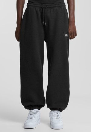 Person, die schwarze elastische Sweatpants mit weißem Logo am Oberschenkel und weiße Sneaker trägt, vor einfachem Hintergrund stehend.