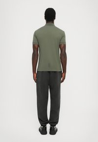 Olive green polo shirt with a fitted design, short sleeves, and a subtle back logo detail, paired with dark, loose-fitting pants and black shoes.