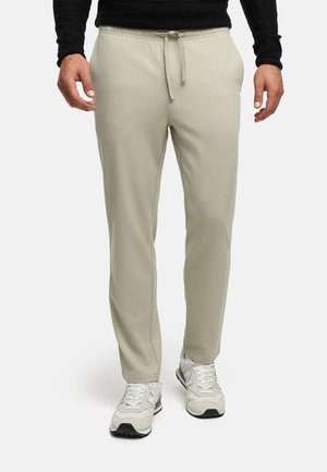 Homme portant un pantalon beige à cordon et des baskets blanches, debout devant un fond clair uni.