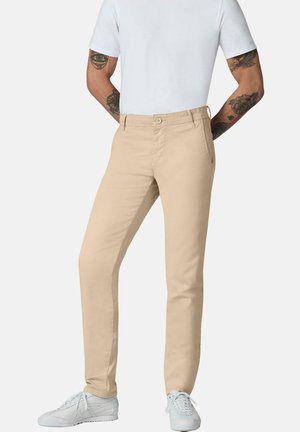 Mann trägt beige slimfit Hose, weiße Sneakers und ein schlichtes weißes kurzärmeliges T-Shirt mit sichtbaren tätowierten Armen.