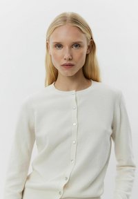 Hvid strikket cardigan med rund halsudskæring, lange ærmer og knaplukning med lyse, reflekterende knapper langs fronten.