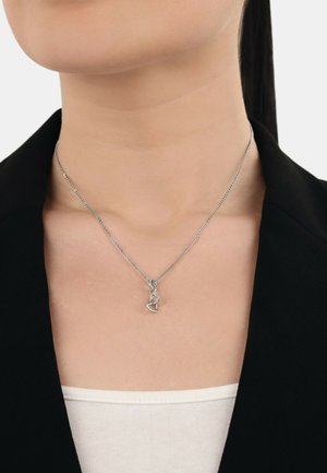 Silberne Halskette mit ineinander verflochtenen Formen und einem kleinen Herzanhänger, an einer feinen Kette. Getragen über einem dunklen Blazer und einem hellen Oberteil.
