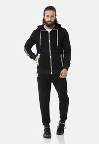 Schwarze Zip-Hoodie und passende Jogginghose, mit einer Kapuze mit Kordelzug, seitlichen Taschen und kontrastierenden Details mit Text.