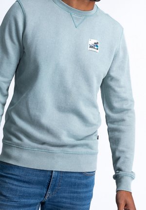 Mand iført lyseblå crewneck hættetrøje med lille grafisk patch på brystet og blå jeans, mod hvid baggrund.