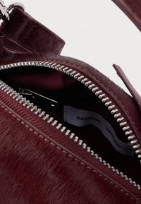 Detailní záběr na otevřenou bordó koženou tašku se stříbrným zipem a viditelnou vnitřní štítkem s nápisem "SAMSØE SAMSØE".