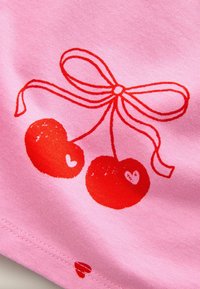 Tela rosa presenta un diseño impreso de dos cerezas rojas con formas de corazón, atadas con un lazo rojo. Costura visible en el dobladillo.