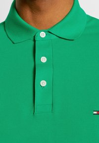 Personne portant un polo vert vif avec trois boutons blancs et un petit logo multicolore rectangulaire sur la poitrine.