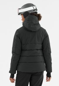 Zwarte pufferjack met een capuchon, voorzien van horizontale stiksels en elastische polsbanden, gecombineerd met een zwarte sk helmet en goggles.