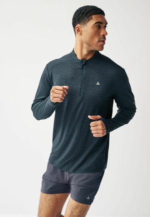 Langärmliges, dunkelgraues Sportoberteil mit einem Viertelreißverschlusskragen, strukturiertem Stoff und Logo auf der Brust, kombiniert mit grauen Shorts.