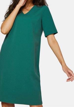 Femme portant une robe bleu sarcelle à manches courtes, longueur genou, col en V, coupe ample, posant une main près du visage et l'autre étendue vers le bas.